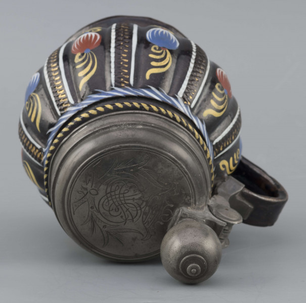 S/CS/595/ML - Kufel  kulisty, o niskiej konicznej stopie i niskiej szyi z pierścieniem, ucho taśmowe w kształcie „C”, pokrywka cynowa. Korpus pionowo kanelowany, podzielony na pasy na przemian gładkie i rytowane w ukośną kratkę. Pokryty szkliwem w kolorze ciemnego kobaltu, zdobiony malowanymi ornamentami podkreślającymi podziały powierzchni, w kolorach: czerwień, żółcień, błękit, biel. Pokrywka płaska, z pierścieniami i uchwytem kulkowym, na zawiasie umocowanym do ucha. Na wierzchu pokrywki grawerunek przedstawiający zawiły ornament – być może monogram spleciony z liter JGL – pod koroną, w uproszczonym wieńcu, z datą 1716.  