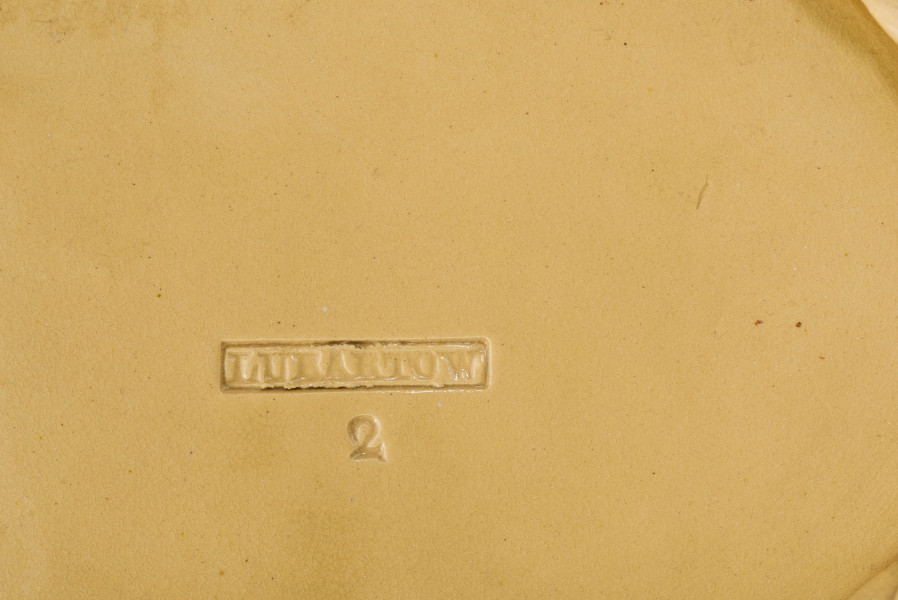 Koszyczek owalny głęboki; dno pełne od zewnątrz obwiedzione warkoczem, boki z okrągłych wałków przeplecionych ukośnie, połączonych profilowaną obręczą tworzącą krawędź. W lustrze (na dnie) dekoracja reliefowa: pośrodku bukiet kwiatów w wazonie, wokół brzegu wić laurowa z owocami. Całość pokryta połyskliwą glazurą w kolorze jasnego ugru.
 