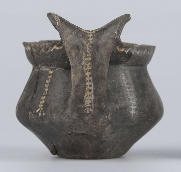 524/A/ML/2 - Kubek gliniany o niskiej szyjce, lejkowato rozchylonej o ukośnie ściętej do wewnątrz krawędzi, oddzielona ostrym załomem od dwustozkowatego brzusca, którego maksymalna wydętość przypada nieco poniżej połowy wysokości naczynia, dno silnie wyodrębnione, wklęsłe. Górny przyczep ucha ansa lunata przylega do krawędzi wylewu, dolny siega największej wydętości brzuśca. Ornament odciśniety prostokątnym stempelkiem, pod wylewem pozioma linia zygzakowata, na brzuścu 6 pionowych pasm ornamentu drabinkowego zakończonych 3 ukośnymi odciskami stempelka (sięgających od załomu szyjka/brzusiec po maksymalną wydętość brzuśca). Takie pasmo także na osi symetrii ucha, pozioma linia drabinkowa łączy skrzydełka ucha anasa lunata. Ornament inkrustowany białą masą. Wymiary: wysokość 7,4 cm; wysokość z uchem ok. 9,1 cm; średnica wylewu 8 cm; szyjka 7,1 cm; brzusiec 9,5 cm; dno 4,4 cm. Surowiec - glina z drobnym piaskiem. Barwa powierzchni zewnętrznej - szarobrunatna, powierzchni wewnętrznej brunatna. Przełom szary. Sposób wykonania - lepienie, powierzchnia gładzona do wybłyszczenia, zdobiona, dobry wypał. zabiegi konserwatorskie - oczyszczanie, sklejanie, wygipsowanie, ucho częściowo zrekonstruowane. Stan zachowania - średni.