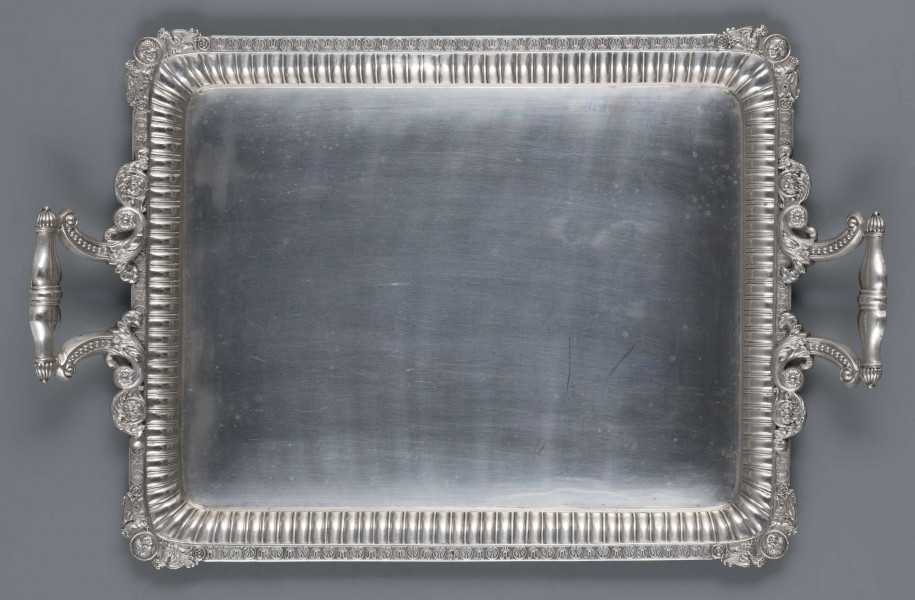 S/Mt/496/ML - Taca prostokątna o zaokrąglonych narożach, z dwoma uchwytami na krótszych bokach. Lustro gładkie, kołnierz podniesiony, trybowany w pionowe paski naprzemian szersze i węższe, brzeg obwiedziony sztancowaną taśmą z motywem palmetek, w narożach wzbogaconym maskami flankowanymi przez gryfy, uchwyty w formie  wolut połączonych toczoną tralką - w osadzeniach liście akantu. 
Na odwrocie sygnatura segmentowa:
- znak miejski Petersburga
- znak imienny złotnika  