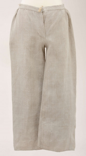 Spodnie lniane, szyte ręcznie z płótna o splocie prostym. Nogawki szerokie proste, u dołu obrębione. Z tyłu zszyte z niewielki trójkątnym karczkiem. U góry ujęte w pasek szer. 3 cm. Zapinane z przodu na 1 guzik wykonany z kawałka drewna. 