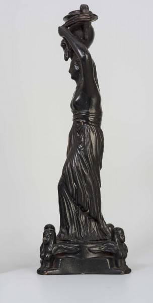 Świecznik figuralny z kariatydą; cokół czworoboczny górą przechodzący w koło, w narożach figurki sfinksów, na nim stojąca postać kobiety z wazonem na głowie, który podtrzymuje uniesionymi rękami. Kobieta do pasa naga, ubrana jest w fałdzistą szatę przwiązaną u góry, na głowie chusta opadajaca na ramiona. Całość malowana lakierem na czarno.
