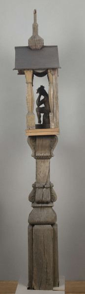 E/8062/1/ML - Kapliczka słupowa wykonana ręcznie siekierą i dłutami. Dębowy, profilowany ozdobnie słup w formie czterograniastej kolumny. Baza przy podstawie spróchniała, ma wgłębione narożniki. Żłobienia, głębokości 3,5 cm, uwypuklają płaszczyzny. Na przedniej ścianie wyryty napis: „Obrobił i postawił własnemi rencznie Jan Oliński dnia 17 marca 1933 r.”. Baza zakończona elementem o przekroju odwróconego trapezu wys. 17 cm i podstawie 26 cm, o zaokrąglonych dolnych narożnikach. Na nim trzon ozdobny czworograniasty, wys. 18 cm. Podstawa trzonu i głowica 4 esowatych żeberek, szerokości 12 cm. Na bocznych pełnych ściankach żeberek uproszony liść akantu. Na głowicach – kapliczka ażurowa. Kolumienki i szczyt brzozowe. Baza kolumny ma kształt graniastosłupa (wys. 12 cm, szer. 7 cm); trzon walcowaty (wys. 22 cm, szer. 8 cm), wybrzuszony, z pionowymi szerokimi rowkowaniami. Głowica (wys. 17 cm), utworzona z uproszczonych liści akantu i dwu złączonych w podstawy rozbieżnych wolut. Szczyt – tympanon ze śladami błękitnej polichromii, ozdobiony perliście małymi sześcianami o zaokrąglonych dolnych krawędziach. U szczytu drewniana kotara (lambrekin) z udrapowanych łukowatych żłobień, w środku ujęty prostą taśmą. Dach dwuspadowy, sosnowy o podciętych i podwiniętych krawędziach dolnych, pokryty blachą. Plecy i podstawa kapliczki – proste, sosnowe.