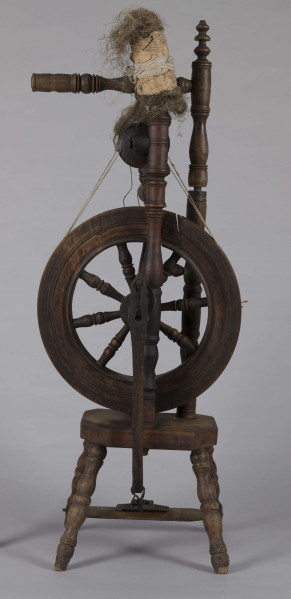 E/16052/ML - Kołowrotek pionowy, drewniany, ozdobnie toczony. Drewno w kolorze ciemnobrązowym. Podstawa to mały stołeczek (wys. 26 cm) o zarysie prostokąta, na 4 toczonych nóżkach o rozstawie u dołu 34 x 30 cm. Pod nim zamocowany podnóżek – pedał do napędzania koła zamachowego, który z ośką koła łączy się poprzez pionowe ramię. W stołeczku 3 pionowe toczone kolumienki. Pomiędzy dwoma z nich umieszczona metalowa ośka, a na niej koło napędowe z ozdobnie toczonymi szprychami. Nad kołem umocowana kolejna pozioma metalowa ośka wraz ze szpulą nawijającą uprzędzione nici. Długość szpuli 29 cm. Trzecia kolumienka służy do zamocowania ramienia z przęślicą (dł. 49 cm) i przędzą. Koło napędowe ze szpulą łączy linka z lnianego sznurka założona na obwód koła i szpuli.