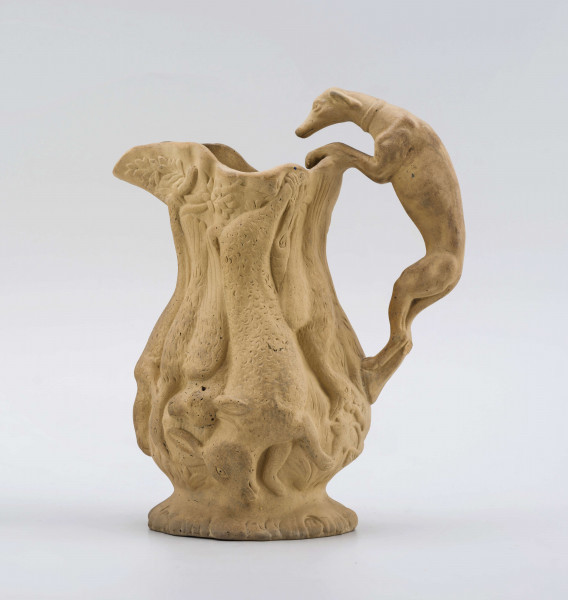 Dzbanek o kształcie zbliżonym do tralki na niskiej szerokiej stopie, z uchem; opracowany reliefowo sprawia wrażenie wydrążonego pnia drzewa, na którym zawieszone są upolowane ptaki i zwierzęta (m. in. bażanty, kaczki, zające, lis), ucho tworzy pełnoplastyczna figurka charta wspinającego się na wyciągniętych łapach i zaglądającego do środka. Powierzchnia matowa, bez polewy.
Na spodzie sygnatura odciśnięta w masie:
- Lubartów – kursywą
