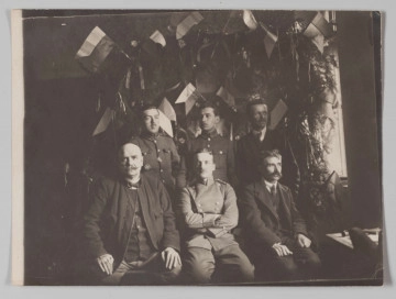 MPol/679/ML MPol/679/ML - Fotografia przedstawia grupę siedmiu mężczyzn: trzech stojących i czterech siedzących przed nimi. Po środku siedzi Jerzy Pol w mundurze wojskowym. Tło stanowi ściana udekorowana polskimi flagami i zielonymi gałązkami.