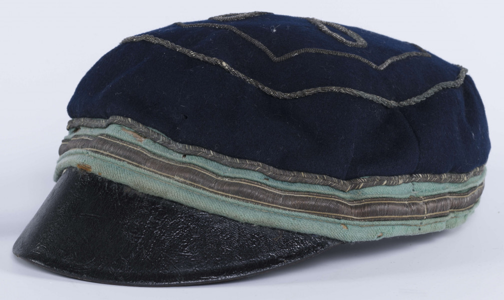 ML/H/2208 - Czapka, czyli tzw. dekiel członka Polskiej Akademickiej Korporacji Astrea działającej na Katolickim Uniwersytecie Lubelskim w latach 1923-1939. Nakrycie głowy w barwach korporanckich, uszyte z nieusztywnianego granatowego sukna, z daszkiem z czarnej skóry, z otokiem w kolorach błękitnym i srebrnym. Na zewnętrznej stronie denka w centralnej części wyszyty fabrycznie srebrną nicią znak korporacji (stylizowany na gwiazdę, w postaci połączonych ze sobą ozdobnych czterech liter „A”) umieszczony wewnątrz haftowanego oktogramu otoczonego kołem z naszytego ozdobnego sznureczka. Po bokach widoczne miejsca zszycia materiału. Wewnątrz podszewka w kolorze granatowym oraz wzmacniający skórzany pasek w kolorze brązowym naokoło.