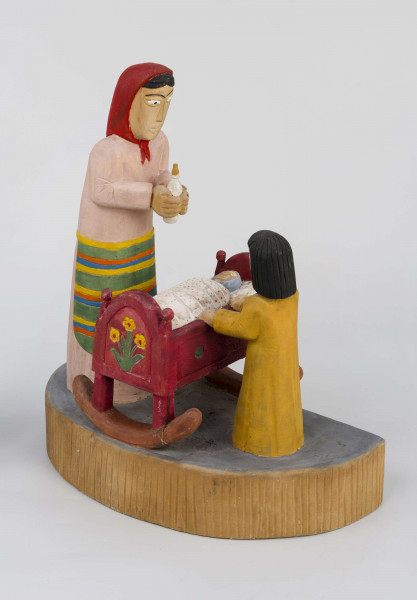 Rzeźba w drewnie lipowym, polichromowana, przedstawiająca postacie matki i dziecka przy kołysce z niemowlęciem. Całość zakomponowana na postumencie wys. 4,4 cm i dł. 22 cm, o kształcie z przodu półokrągłym. Pośrodku usytuowana kołyska na biegunach, wys. 9 cm i dł. 11,2 cm. Kołyska czerwona, ścianki szczytowe ozdobione bukietami składającymi się z trzech wielopłatkowych kwiatów w kolorze żółtym i tulipanowych zielonych liśćmi. W kołysce, w beciku białym, w czerwono-zielone kropki, dziecko, w niebieskiej chustce na głowie. Z lewej strony kołyski postać matki, w długiej jasnoróżowej sukni, w pasie przepasanej fartuchem w żółto-zielono-pomarańczowo-niebieskie poziome pasy. Twarz płaska, nos długi prosty, usta wąskie małe, oczy owalne o podkreślonych białych białkach i czarnych źrenicach, brwi łukowate czarne. Na głowie czerwona chustka zawiązana pod brodą. Spod niej wystają czarne włosy. W rękach trzyma butelkę z mlekiem. Z prawej strony kołyski stoi dziecko w żółtej sukience z niebieskim kołnierzykiem. Twarz dziecka płaska, nos prosty wąski, usta małe, oczy owalne, brwi proste czarne. Włosy czarne, proste sięgające do ramion, zaczesane z grzywką na czole. Ręce wsparte na kołysce. 