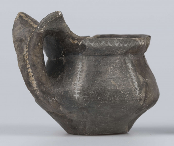 524/A/ML/2 - Kubek gliniany o niskiej szyjce, lejkowato rozchylonej o ukośnie ściętej do wewnątrz krawędzi, oddzielona ostrym załomem od dwustozkowatego brzusca, którego maksymalna wydętość przypada nieco poniżej połowy wysokości naczynia, dno silnie wyodrębnione, wklęsłe. Górny przyczep ucha ansa lunata przylega do krawędzi wylewu, dolny siega największej wydętości brzuśca. Ornament odciśniety prostokątnym stempelkiem, pod wylewem pozioma linia zygzakowata, na brzuścu 6 pionowych pasm ornamentu drabinkowego zakończonych 3 ukośnymi odciskami stempelka (sięgających od załomu szyjka/brzusiec po maksymalną wydętość brzuśca). Takie pasmo także na osi symetrii ucha, pozioma linia drabinkowa łączy skrzydełka ucha anasa lunata. Ornament inkrustowany białą masą. Wymiary: wysokość 7,4 cm; wysokość z uchem ok. 9,1 cm; średnica wylewu 8 cm; szyjka 7,1 cm; brzusiec 9,5 cm; dno 4,4 cm. Surowiec - glina z drobnym piaskiem. Barwa powierzchni zewnętrznej - szarobrunatna, powierzchni wewnętrznej brunatna. Przełom szary. Sposób wykonania - lepienie, powierzchnia gładzona do wybłyszczenia, zdobiona, dobry wypał. zabiegi konserwatorskie - oczyszczanie, sklejanie, wygipsowanie, ucho częściowo zrekonstruowane. Stan zachowania - średni.