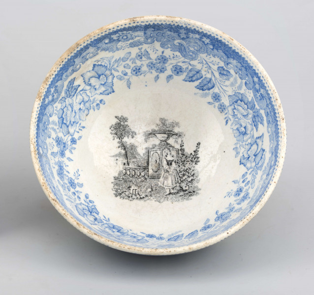 Miseczka półkulista, na niskiej, okrągłej stopce. Zdobiona od strony zewnetrznej i wewnętrznej nadrukiem w kolorze błękitnym z wzorem wolut i ukwieconych gałązek róży i goździków. W lustrze scena w kolorze czarnym przedstawiająca młodą dziewczynę w ogrodzie z elementami 