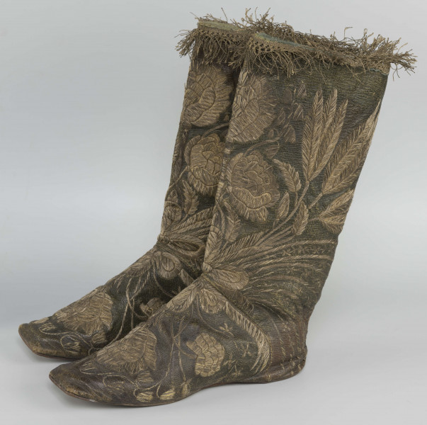 S/T/42/ML - Para butów z wysokimi cholewkami. Cholewka z miękkiej,zielonej skóry,  z dekoracyjnym haftem złotą nicią. Haft z dużym motywem kwiatowym: piwonie, kampanule i drobne kwiatki niezapominajki, tulipany(?), wydłużone liście i wici roślinne, wygięte łukowato.  Wewnatrz buty mają wykończenie białym, cienkim płótnem. Brzegi cholewki obszyte złota koronką z frędzlą. Podeszwy cieńkie z jednego kawałka skóry, bez obcasów, płaskie, z przodu prosto ścięte. Obydwie, skórzane podeszwy mają taki sam wykrój, bez rozróżnienia na lewą i prawą.