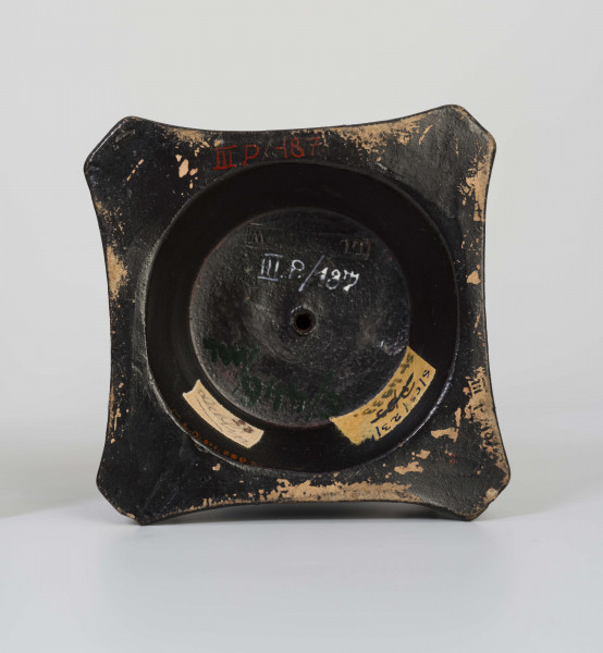 Świecznik figuralny z kariatydą; cokół czworoboczny górą przechodzący w koło, w narożach figurki sfinksów, na nim stojąca postać kobiety z wazonem na głowie, który podtrzymuje uniesionymi rękami. Kobieta do pasa naga, ubrana jest w fałdzistą szatę przwiązaną u góry, na głowie chusta opadajaca na ramiona. Całość malowana lakierem na czarno.
