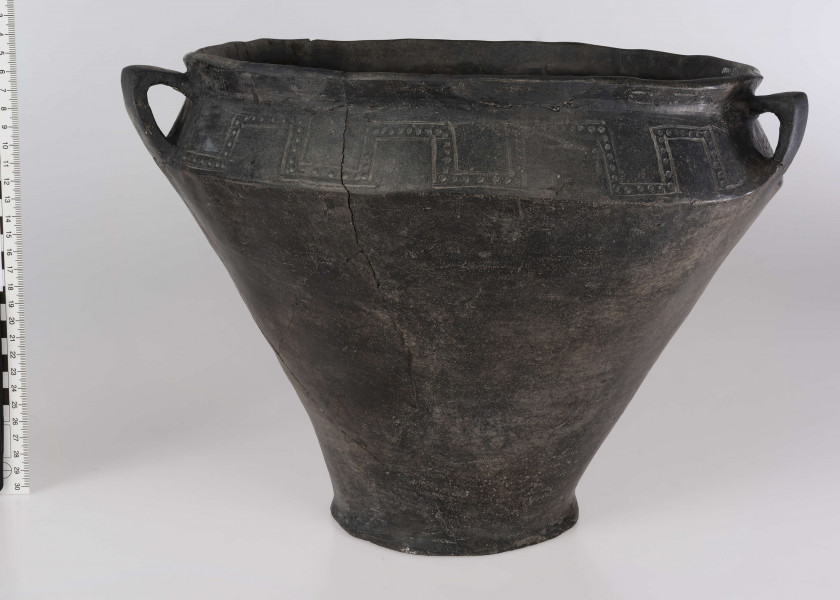 765/A/ML/1 - Waza wyraźnie profilowana, z prostym, odgietym na zewnatrz brzegiem, ostro zaznaczonym załomem brzuśca i wyodrębnionym dnem. Ponad załomem brzuśca występuje ornament w postaci: jodełek, swastyk, tworzonych przez dwie linie ryte i rząd dołków pomiędzy nimi oraz ornament blankowy. Naczynie ma trzy taśmowate ucha kolankowato zgięte. 
