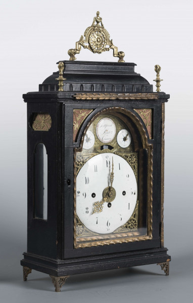 S/Mt/409/ML - Obudowa szafkowa, czworoboczna ze schodkowym zwieńczeniem, pokryta czarną politurą, oszklona z czterech stron, na czterech ażurowych nóżkach z mosiądzu; mosiężny uchwyt, ozdobny, składany a w górnych narożnikach sterczyny w formie płonących wazonów. Mechanizm chodu i bicia o napędzie sprężynowym, z wahadłem i dwoma dzwonkami; bicie godzin i kwadransów, budzik i repetier. Tarcza biało emaliowana z czarnymi cyframi arabskimi godzin i oznaczeniem minut, w niej trzy otwory na osie sprężyn; dwie ozdobne wskazówki. Powyżej tarczy wycięcie z widocznym wahadełkiem kontrolnym i trzy mniejsze emaliowane tarcze: środkowa z sygnaturą: Gottfried / Krohz / Cracovie, boczne z wyłączeniem chodu i bicia.