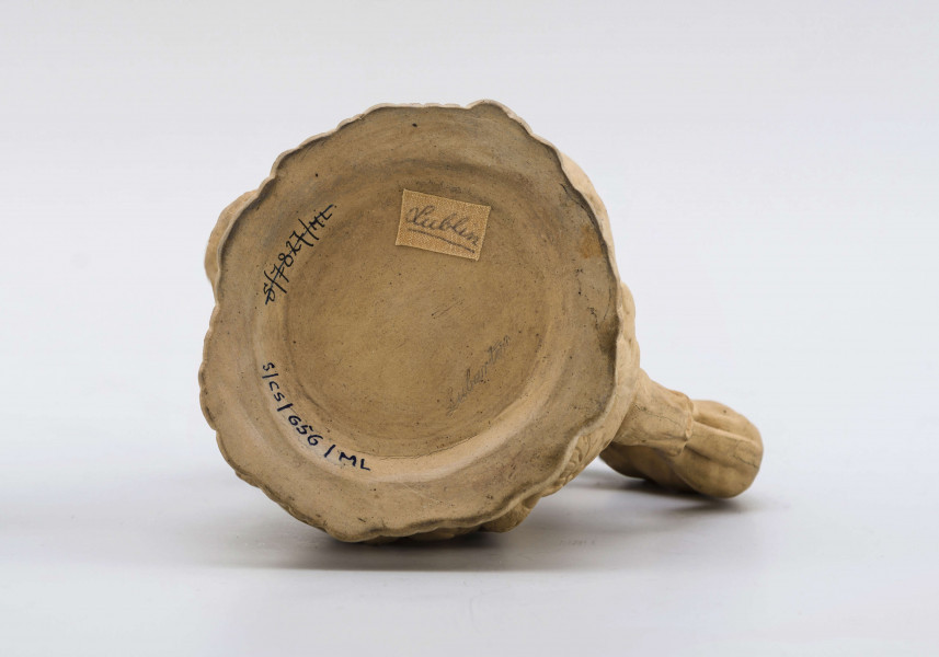 Dzbanek o kształcie zbliżonym do tralki na niskiej szerokiej stopie, z uchem; opracowany reliefowo sprawia wrażenie wydrążonego pnia drzewa, na którym zawieszone są upolowane ptaki i zwierzęta (m. in. bażanty, kaczki, zające, lis), ucho tworzy pełnoplastyczna figurka charta wspinającego się na wyciągniętych łapach i zaglądającego do środka. Powierzchnia matowa, bez polewy.
Na spodzie sygnatura odciśnięta w masie:
- Lubartów – kursywą
