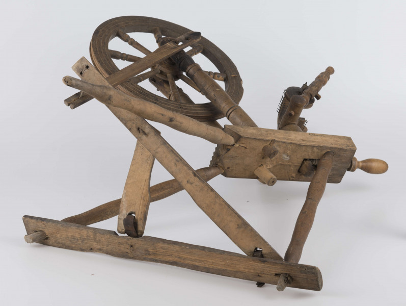 E/16647/ML - Kołowrotek poziomy, drewniany, ozdobnie toczony. Drewno niemalowane. Podstawa to 3 skośnie ustawione nóżki. Dwie z nich osadzone w wąskiej listwie. Górne końce nóżek osadzone w prostokątnej pochyłej grubej desce (11 x 32 x 4 cm). Stanowi ona podstawę do osadzenia koła napędowego i szpuli zwijającej nici. W niej zamocowany uchwyt połączony ze śrubą, która reguluje położenie szpuli. Koło osadzone na metalowej ośce posiada 8 toczonych ozdobnie szprych. Jeden koniec ośki połączony pionową ruchomą listewką z pedałem wprawiającym w ruch kołowrotek. Szpula osadzona również na metalowej ośce.