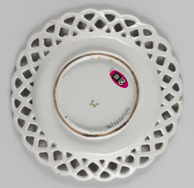 S/CS/1523/ML - Talerz płytki z szerokim ażurowym kołnierzem ze skośnej kratownicy o falistym konturze brzegu. Kratownica zdobiona na złączach kwiatkami. Pokryty polewą w kolorze kremowobiałym; w lustrze duża jasnozielona gałązka z kawiatami w kolorze fioletowej purpury, tego samego koloru kwiatki na kołnierzu.
 