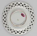 S/CS/1523/ML - Talerz płytki z szerokim ażurowym kołnierzem ze skośnej kratownicy o falistym konturze brzegu. Kratownica zdobiona na złączach kwiatkami. Pokryty polewą w kolorze kremowobiałym; w lustrze duża jasnozielona gałązka z kawiatami w kolorze fioletowej purpury, tego samego koloru kwiatki na kołnierzu.
 