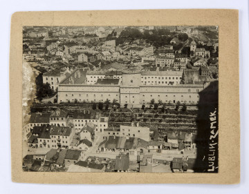 ML/H/F/238 Fotografia czarnobiała (widok z lotu ptaka) przedstawiająca Wzgórze Zamkowe i jego okolice. Ujęcie od strony Wzgórza Staromiejskiego. W centralnej części zabudowania więzienia na zamku, z zabytkową Kaplicą Trójcy Świętej oraz donżonem. Przed gmachem, po lewej, budynek administracji więziennej (rozebrany w 1953 r.). U podnóża zamku dzielnica żydowska z zabudową ulic: Krawieckiej (Dom Modlitwy), Zamkowej, Jatecznej (Wielka Synagoga / Synagoga Maharszala), Nadstawnej, Ruskiej (cerkiew prawosławna), Czwartek oraz Lubartowskiej. Wzdłuż prawej krawędzi na zaciemnionym tle skrzydła samolotu napis białymi literami naniesiony na fotografię. Zdjęcie podklejone na tekturę.