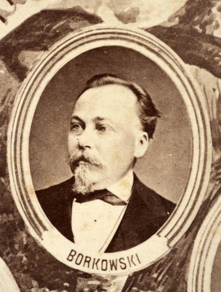 Fotografie w odcieniu sepii umieszczone na tle drzew i roślin, w centralnym miejscu postać kobiety ze sztandarem i aniołkami, wkomponowane widoki budowanej stacji, mostu kolejowego i lokomotywy. Największe zdjęcie w górnej, środkowej części bez podpisu, prawdopodobnie inż. głównej kolei nadwiślańskiej Tadeusz Chrzanowski lub jego następca Walery Titkow. Wśród podobizn pracownicy Biura Głównego Inżyniera: inż. Włodzimierz Stanisławski - pomocnik głównego inżyniera; inż. Feliks Kucharzewski -  pomocnik naczelnika biura; Feliks Kamocki - naczelnik kancelarii; Witold Lanci - naczelnik Wydz. Architektury; dr Wacław Lasocki- służba zdrowia; Władysław Radwan - sprawy zakupu gruntów; inż. Stanisław hr Scipio - projektant mostów kolejowych i jego współpracownicy: inż. Stefan Zieliński, inż. Maurycy Machalski, inż. Feliks Kucharzewski. Fotografia w passe - partout, za szkłem, oprawiona w drewnianą, profilowaną ramę.