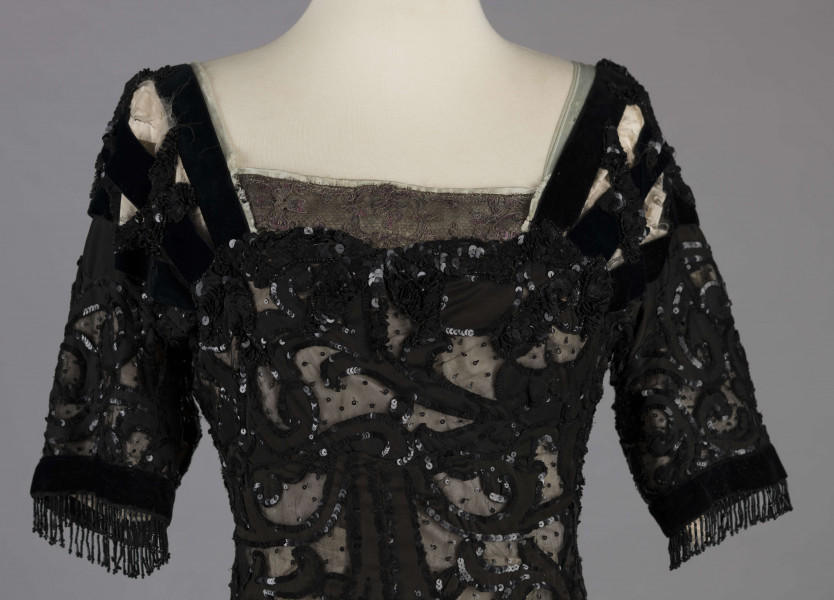 S/T/243/ML - Suknia wizytowa, czarna, uszyta z atłasu jedwabnego, jedwabnego tiulu i aksamitu, haftowana cekinami. Krótki rękaw z jedwabnego czarnego aksamitu, dekolt prostokatny, dość głęboki, wykończony wstawką haftowaną i aksamitem. Kolory są kontrastowe. Spodnia warstwa sukni, z białej tafty i muślinu, widoczna w partiach sukni szytych z czarnego tiulu, dodaje całości elegancji. Brzegi dekoracyjnej wici z tafty zaakcentowane naszytymi cekinami. Suknia do ziemi z tyłu krótki tren. Brak halki. 