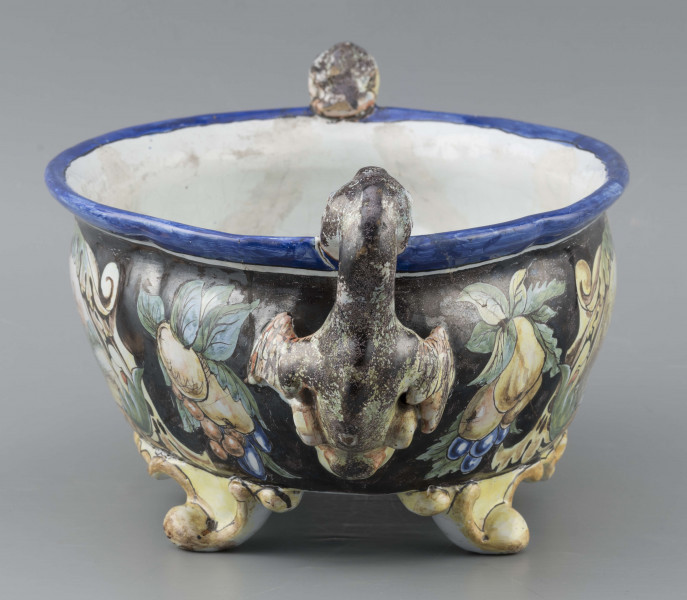 S/CS/90/ML - Żardiniera owalna, na czterech konsolkowych nóżkach połączonych falistymi spływami, wylew z lekko rozchylonym brzegiem, po bokach dwa uchwyty w formie uskrzydlonych smoków, pionowo osadzone. Powierzchni malowana wielobarwnie:  po obu stronach bogate kartusze wolutowo-liściaste z pejzażami  -  jeden z zamkiem obronnym, drugi z wierzbą, po bokach kartuszy festony owoców na ciemno granatowym tle. Wylew obwiedziony błękitnym pasem, wnętrze białobłękitne. 