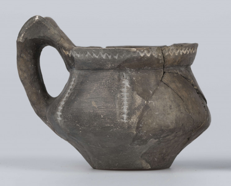 524/A/ML/2 - Kubek gliniany o niskiej szyjce, lejkowato rozchylonej o ukośnie ściętej do wewnątrz krawędzi, oddzielona ostrym załomem od dwustozkowatego brzusca, którego maksymalna wydętość przypada nieco poniżej połowy wysokości naczynia, dno silnie wyodrębnione, wklęsłe. Górny przyczep ucha ansa lunata przylega do krawędzi wylewu, dolny siega największej wydętości brzuśca. Ornament odciśniety prostokątnym stempelkiem, pod wylewem pozioma linia zygzakowata, na brzuścu 6 pionowych pasm ornamentu drabinkowego zakończonych 3 ukośnymi odciskami stempelka (sięgających od załomu szyjka/brzusiec po maksymalną wydętość brzuśca). Takie pasmo także na osi symetrii ucha, pozioma linia drabinkowa łączy skrzydełka ucha anasa lunata. Ornament inkrustowany białą masą. Wymiary: wysokość 7,4 cm; wysokość z uchem ok. 9,1 cm; średnica wylewu 8 cm; szyjka 7,1 cm; brzusiec 9,5 cm; dno 4,4 cm. Surowiec - glina z drobnym piaskiem. Barwa powierzchni zewnętrznej - szarobrunatna, powierzchni wewnętrznej brunatna. Przełom szary. Sposób wykonania - lepienie, powierzchnia gładzona do wybłyszczenia, zdobiona, dobry wypał. zabiegi konserwatorskie - oczyszczanie, sklejanie, wygipsowanie, ucho częściowo zrekonstruowane. Stan zachowania - średni.