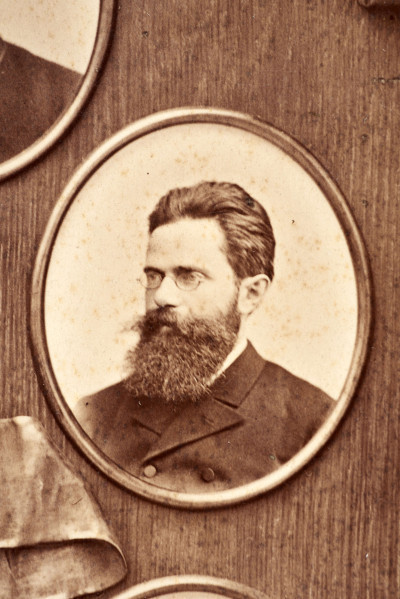 Pośrodku prostokątnego pola o zaokrąglonych narożnikach większa fotografia przyozdobiona wstęgą i gałązkami wawrzynu. Dookoła szesnaście symetrycznie rozmieszczonych mniejszych fotografii. W prawym górnym rogu dr Wacław Lasocki, naczelny lekarz kolei nadwiślańskiej w latach 1875 - 78. W polu ramy w górnej części medalion z płaskorzeźbionym popiersiem z profilu. Przedstawia Leopolda Kronenberga - ojca. Po bokach dwia półleżące aniołki trzymają wieniec laurowy. Po lewej stronie ramy postać robotnika Poleszuka, po prawej pracownika warsztatów kolejowych. W dolnej części front lokomotywy i złoty żeton uprawniający do bezpłatnej jazdy pociągiem I klasy. Fotografia naklejona na tekturę, w dolnej części u dołu sucha pieczęć zakładu fotograficznego.