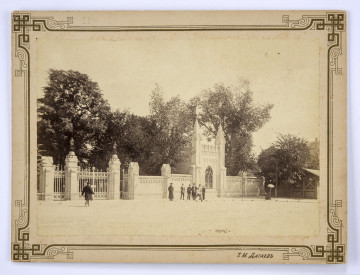 Wejście do Ogrodu Saskiego w Lublinie. Ogrodzenie murowane w stylu neogotyckim. Przed nim stoi sześciu mężczyzn w różnym wieku. Pierwszy z lewej to prawdopodobnie stróż w długim płaszczu i czapce z daszkiem. Po lewej stronie widać maszerującego mężczyznę ubranego w kurtkę i czapkę. Po prawej stoi kobieta w ciemnej sukni z rozłożoną nad głową parasolką. W linii muru wyróżnia się fasada Domku Odźwiernego ozdobionego sterczynami, attyką i ostrołukowym oknem. Po lewej stronie brama główna i kute w metalu dwie furtki prowadzące na teren ogrodu. Na murowanych słupkach ustawione latarnie. Po prawej stronie widoczny wolnostojący mały parterowy budynek ze spadzistym dachem oraz szczyt kamienicy (częściowo przysłonięte zielenią). Za ogrodzeniem wysokie drzewa parku. Od strony ulicy widoczna brukowana nawierzchnia. 
Fotografia sepiowana, naklejona na tekturę, otoczona ozdobną ramką w kolorze złotym o motywach geometryczno-roślinnych. W dolnej części, po prawej stronie nazwisko autora fotografii w języku rosyjskim.
