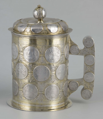 S/Mt/286/ML - Kufel cylindryczny z nakładaną pokrywką, na zewnątrz i wewnątrz złocony (silnie przetarte); dolna krawędź obwiedziona płaską kryzą, analogiczna kryza przy krawędzi pokrywki; pokrywka wysklepiona z kulistym uchwytem; ucho o fantazyjnym kształcie na bazie prostokata. Całość naczynia - płaszcz, dno, pokrywka wraz z uchwytem, ucho - zdobione wmontowanymi srebrnymi monetami. Monety bite w latach 1689 - 1697 przez książąt niemieckich. Na płaszczu trzy rzędy po osiem monet ułożonych naprzemiennie, o wartościach: 3/4 talara, 16 gute grosche i 24 mariengrosche; na pokrywce 10 monet wartości 6 mariengrosche i 3 dalsze na uchwycie;  na uchu takier same po sześć z obu stron; na dnie pośrodku jedna moneta - 24 mariengrosche. Monety płączone rytowanymi poziomymi paskami  z ornamentem akantowym. 
