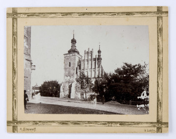 ML/H/F/25/17 Widok zachodniej fasady kościoła pw. Najświętszej Marii Panny Zwycięskiej (tzw. pobrygidkowskiego lub powizytkowskiego) przy ul. Narutowicza (dawniej Namiestnikowskiej). Na pierwszym planie brukowana ulica, częściowo zacieniona. Widać stojącego na niej mężczyznę. Po lewej stronie narożnik ceglanej kamienicy i postać w cieniu. Fasada kościoła przysłonięta drzewem. Czworokątna wieża zwieńczona kopułą oraz trójkątny szczyt widoczne od ulicy. Po obu stronach świątyni za ogrodzeniem drzewa. W głębi wieża Bramy Krakowskiej.
Fotografia sepiowana, naklejona na tekturę, otoczona ozdobną ramką w kolorze złotym w formie przecinających się na rogach listew. W l. d. rogu nazwisko autora fotografii, w p. d. nazwa miasta. Na rewersie odbitki pieczęci tuszowych Muzeum Nałęczowskiego (okrągła, w kolorze fioletowym) oraz Biblioteki Nałęczowskiej (podłużna, w kolorze fioletowym). Wzdłuż górnej krawędzi odręczny napis czarnym tuszem.