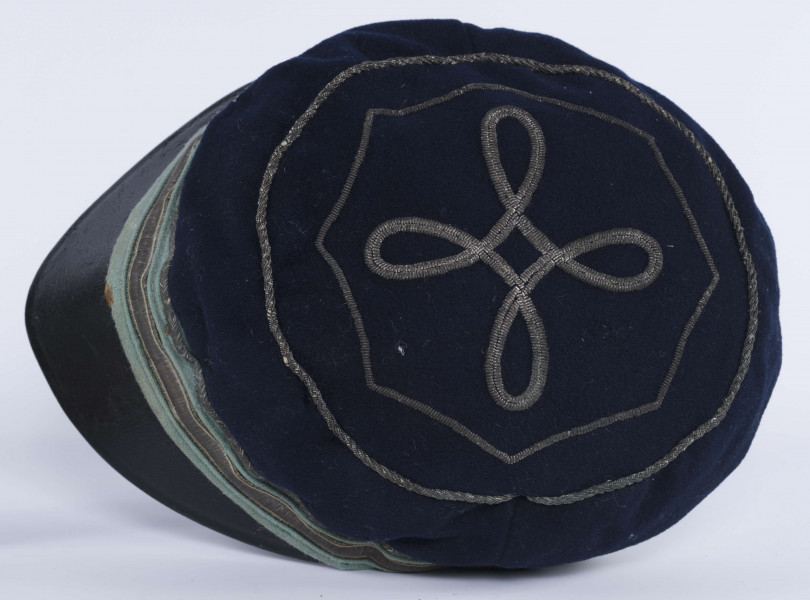 ML/H/2208 - Czapka, czyli tzw. dekiel członka Polskiej Akademickiej Korporacji Astrea działającej na Katolickim Uniwersytecie Lubelskim w latach 1923-1939. Nakrycie głowy w barwach korporanckich, uszyte z nieusztywnianego granatowego sukna, z daszkiem z czarnej skóry, z otokiem w kolorach błękitnym i srebrnym. Na zewnętrznej stronie denka w centralnej części wyszyty fabrycznie srebrną nicią znak korporacji (stylizowany na gwiazdę, w postaci połączonych ze sobą ozdobnych czterech liter „A”) umieszczony wewnątrz haftowanego oktogramu otoczonego kołem z naszytego ozdobnego sznureczka. Po bokach widoczne miejsca zszycia materiału. Wewnątrz podszewka w kolorze granatowym oraz wzmacniający skórzany pasek w kolorze brązowym naokoło.