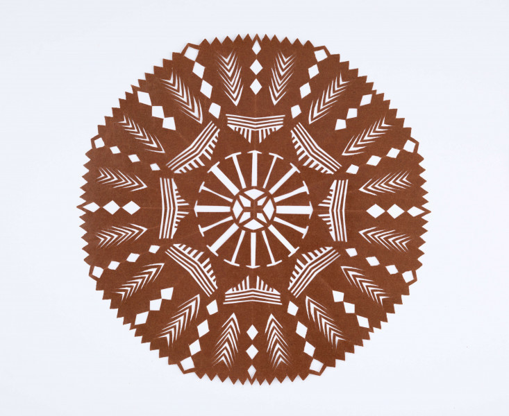 Wycinanka kolista z glansowanego papieru w kolorze brązowym, o ośmiu osiach symetrii. W centrum gwiazda czteroramienna. Na pozostałej powierzchni wycięcia w postaci paseczków pionowych i poziomych, ułożonych grupami. Na otoku wycięcia w postaci rombów i kłosów stylizowanych. 