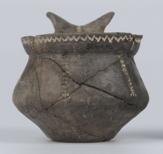 524/A/ML/2 - Kubek gliniany o niskiej szyjce, lejkowato rozchylonej o ukośnie ściętej do wewnątrz krawędzi, oddzielona ostrym załomem od dwustozkowatego brzusca, którego maksymalna wydętość przypada nieco poniżej połowy wysokości naczynia, dno silnie wyodrębnione, wklęsłe. Górny przyczep ucha ansa lunata przylega do krawędzi wylewu, dolny siega największej wydętości brzuśca. Ornament odciśniety prostokątnym stempelkiem, pod wylewem pozioma linia zygzakowata, na brzuścu 6 pionowych pasm ornamentu drabinkowego zakończonych 3 ukośnymi odciskami stempelka (sięgających od załomu szyjka/brzusiec po maksymalną wydętość brzuśca). Takie pasmo także na osi symetrii ucha, pozioma linia drabinkowa łączy skrzydełka ucha anasa lunata. Ornament inkrustowany białą masą. Wymiary: wysokość 7,4 cm; wysokość z uchem ok. 9,1 cm; średnica wylewu 8 cm; szyjka 7,1 cm; brzusiec 9,5 cm; dno 4,4 cm. Surowiec - glina z drobnym piaskiem. Barwa powierzchni zewnętrznej - szarobrunatna, powierzchni wewnętrznej brunatna. Przełom szary. Sposób wykonania - lepienie, powierzchnia gładzona do wybłyszczenia, zdobiona, dobry wypał. zabiegi konserwatorskie - oczyszczanie, sklejanie, wygipsowanie, ucho częściowo zrekonstruowane. Stan zachowania - średni.