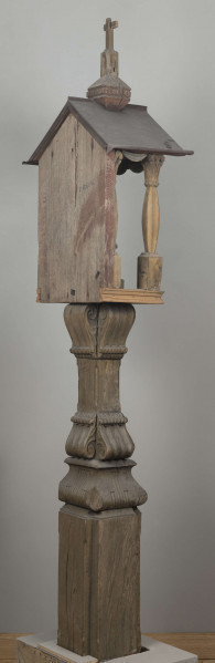 E/8062/1/ML - Kapliczka słupowa wykonana ręcznie siekierą i dłutami. Dębowy, profilowany ozdobnie słup w formie czterograniastej kolumny. Baza przy podstawie spróchniała, ma wgłębione narożniki. Żłobienia, głębokości 3,5 cm, uwypuklają płaszczyzny. Na przedniej ścianie wyryty napis: „Obrobił i postawił własnemi rencznie Jan Oliński dnia 17 marca 1933 r.”. Baza zakończona elementem o przekroju odwróconego trapezu wys. 17 cm i podstawie 26 cm, o zaokrąglonych dolnych narożnikach. Na nim trzon ozdobny czworograniasty, wys. 18 cm. Podstawa trzonu i głowica 4 esowatych żeberek, szerokości 12 cm. Na bocznych pełnych ściankach żeberek uproszony liść akantu. Na głowicach – kapliczka ażurowa. Kolumienki i szczyt brzozowe. Baza kolumny ma kształt graniastosłupa (wys. 12 cm, szer. 7 cm); trzon walcowaty (wys. 22 cm, szer. 8 cm), wybrzuszony, z pionowymi szerokimi rowkowaniami. Głowica (wys. 17 cm), utworzona z uproszczonych liści akantu i dwu złączonych w podstawy rozbieżnych wolut. Szczyt – tympanon ze śladami błękitnej polichromii, ozdobiony perliście małymi sześcianami o zaokrąglonych dolnych krawędziach. U szczytu drewniana kotara (lambrekin) z udrapowanych łukowatych żłobień, w środku ujęty prostą taśmą. Dach dwuspadowy, sosnowy o podciętych i podwiniętych krawędziach dolnych, pokryty blachą. Plecy i podstawa kapliczki – proste, sosnowe.