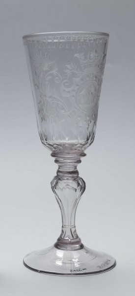 S/CS/244/ML - Stopa okrągła, lekko uniesiona, trzon tralkowy, z pęcherzykiem powietrza, fasetowany, od czaszy oddzielony pierścieniem i dwoma talerzykami, czasza stozkowata, u nasady zaokrąglona, szlifowana w dwa pionowe rzędy owalnych faset. Dekoracja rytowana: na czaszy dwustronna - z jednej strony owalna tarcza z parą gołąbków połączonych dziobkami stojących na serduszkach w rozbudowanym kartuszu z uszakami, z którego wyrastaja gałązki; po drugiej stronie litery 