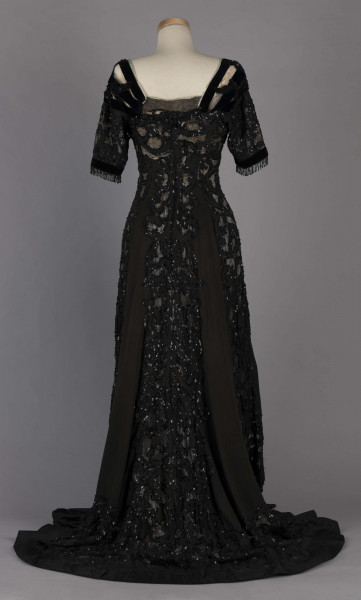 S/T/243/ML - Suknia wizytowa, czarna, uszyta z atłasu jedwabnego, jedwabnego tiulu i aksamitu, haftowana cekinami. Krótki rękaw z jedwabnego czarnego aksamitu, dekolt prostokatny, dość głęboki, wykończony wstawką haftowaną i aksamitem. Kolory są kontrastowe. Spodnia warstwa sukni, z białej tafty i muślinu, widoczna w partiach sukni szytych z czarnego tiulu, dodaje całości elegancji. Brzegi dekoracyjnej wici z tafty zaakcentowane naszytymi cekinami. Suknia do ziemi z tyłu krótki tren. Brak halki. 