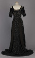 S/T/243/ML - Suknia wizytowa, czarna, uszyta z atłasu jedwabnego, jedwabnego tiulu i aksamitu, haftowana cekinami. Krótki rękaw z jedwabnego czarnego aksamitu, dekolt prostokatny, dość głęboki, wykończony wstawką haftowaną i aksamitem. Kolory są kontrastowe. Spodnia warstwa sukni, z białej tafty i muślinu, widoczna w partiach sukni szytych z czarnego tiulu, dodaje całości elegancji. Brzegi dekoracyjnej wici z tafty zaakcentowane naszytymi cekinami. Suknia do ziemi z tyłu krótki tren. Brak halki. 