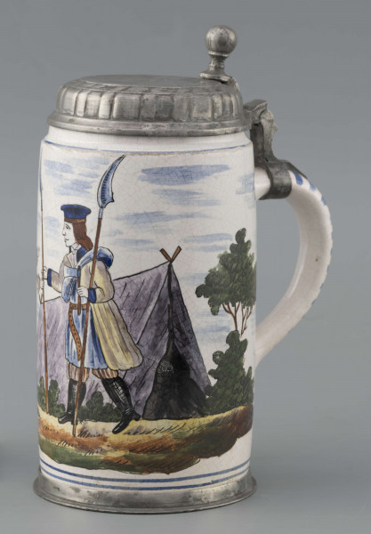 S/CS/64/ML - Korpus cylindryczny z uchem w kształcie litery „C”, pokrywka cynowa, płaska z profilowanym brzegiem i kulistą sterczyną  na zawiasie umocowanym do ucha; profilowana cynowa obręcz otacza dolną krawędź. Pokryty białym szkliwem z wielobarwną dekoracją malowaną skomponowaną przeciwlegle do ucha. Przedstawia ona w uproszczony sposób scenę w obozie  wojskowym: dwaj uzbrojeni kosynierzy stoją na wprost siebie, za nimi widoczny namiot a wokół zielone zarośla i drzewa. Niebo zaznaczone błękitnymi plamami, w tym samym kolorze malowane paski obiegają krawędzie naczynia i zdobią ucho. Tematyka przedstawienia ma wyraźnie patriotyczny charakter nawiązując do powstania kościuszkowskiego.

