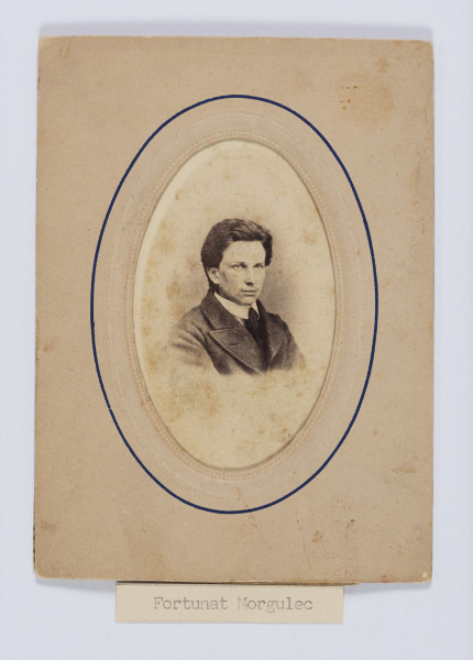 ML/H/F/72 - Fotografia przedstawia młodego człowieka w popiersiu, zwróconego lekko w lewo. Twarz młoda, rysy regularne. Włosy ciemne, gęste, zaczesane od góry w bok. Ubrany w białą koszulę, krawat, kurtke z szerokimi klapami. 
Na kopertce passepartout z tyłu wypisane czarnym atramentem innym charakterem pisma - Fortunat Morgulec.