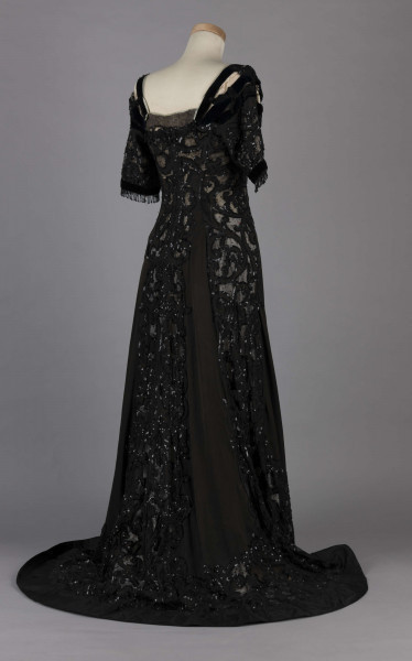 S/T/243/ML - Suknia wizytowa, czarna, uszyta z atłasu jedwabnego, jedwabnego tiulu i aksamitu, haftowana cekinami. Krótki rękaw z jedwabnego czarnego aksamitu, dekolt prostokatny, dość głęboki, wykończony wstawką haftowaną i aksamitem. Kolory są kontrastowe. Spodnia warstwa sukni, z białej tafty i muślinu, widoczna w partiach sukni szytych z czarnego tiulu, dodaje całości elegancji. Brzegi dekoracyjnej wici z tafty zaakcentowane naszytymi cekinami. Suknia do ziemi z tyłu krótki tren. Brak halki. 