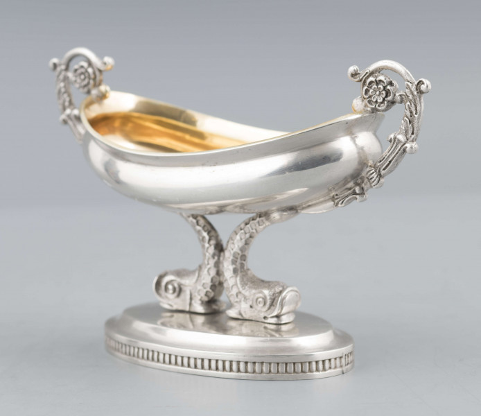 S/Mt/511/ML - Solniczka w formie łódki wspartej na dwóch delfinach, z owalną podstawą, z dwoma uchami z wici roślinnej. Brzeg czarki wywinięty, podstawa lekko podniesiona a jej brzeg zdobiony pionowym karbowaniem. Na spodzie sygnatura:
- imienna 