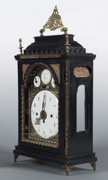 S/Mt/409/ML - Obudowa szafkowa, czworoboczna ze schodkowym zwieńczeniem, pokryta czarną politurą, oszklona z czterech stron, na czterech ażurowych nóżkach z mosiądzu; mosiężny uchwyt, ozdobny, składany a w górnych narożnikach sterczyny w formie płonących wazonów. Mechanizm chodu i bicia o napędzie sprężynowym, z wahadłem i dwoma dzwonkami; bicie godzin i kwadransów, budzik i repetier. Tarcza biało emaliowana z czarnymi cyframi arabskimi godzin i oznaczeniem minut, w niej trzy otwory na osie sprężyn; dwie ozdobne wskazówki. Powyżej tarczy wycięcie z widocznym wahadełkiem kontrolnym i trzy mniejsze emaliowane tarcze: środkowa z sygnaturą: Gottfried / Krohz / Cracovie, boczne z wyłączeniem chodu i bicia.