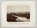 Na pierwszym planie rozlewisko rzeki porośnięte trawą. Dalej most żelazny kratowy długości 165 m, trzy przęsła po 53 m. Na prawym brzegu na nasypie wagony i kilku mężczyzn. Na przeciwległym brzegu widoczne rusztowanie. Fotografia o zaokrąglonych rogach, obrysowana dookoła cienką ramką, naklejona na tekturę. Powyżej i pod fotografią napisy po rosyjsku - z lewej i po polsku - z prawej strony. W dolnej części informacja o autorze zdjęcia po rosyjsku.
