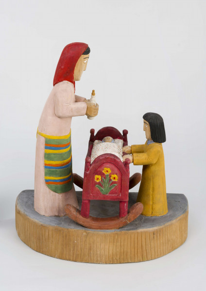 Rzeźba w drewnie lipowym, polichromowana, przedstawiająca postacie matki i dziecka przy kołysce z niemowlęciem. Całość zakomponowana na postumencie wys. 4,4 cm i dł. 22 cm, o kształcie z przodu półokrągłym. Pośrodku usytuowana kołyska na biegunach, wys. 9 cm i dł. 11,2 cm. Kołyska czerwona, ścianki szczytowe ozdobione bukietami składającymi się z trzech wielopłatkowych kwiatów w kolorze żółtym i tulipanowych zielonych liśćmi. W kołysce, w beciku białym, w czerwono-zielone kropki, dziecko, w niebieskiej chustce na głowie. Z lewej strony kołyski postać matki, w długiej jasnoróżowej sukni, w pasie przepasanej fartuchem w żółto-zielono-pomarańczowo-niebieskie poziome pasy. Twarz płaska, nos długi prosty, usta wąskie małe, oczy owalne o podkreślonych białych białkach i czarnych źrenicach, brwi łukowate czarne. Na głowie czerwona chustka zawiązana pod brodą. Spod niej wystają czarne włosy. W rękach trzyma butelkę z mlekiem. Z prawej strony kołyski stoi dziecko w żółtej sukience z niebieskim kołnierzykiem. Twarz dziecka płaska, nos prosty wąski, usta małe, oczy owalne, brwi proste czarne. Włosy czarne, proste sięgające do ramion, zaczesane z grzywką na czole. Ręce wsparte na kołysce. 