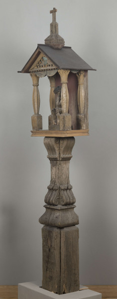 E/8062/1/ML - Kapliczka słupowa wykonana ręcznie siekierą i dłutami. Dębowy, profilowany ozdobnie słup w formie czterograniastej kolumny. Baza przy podstawie spróchniała, ma wgłębione narożniki. Żłobienia, głębokości 3,5 cm, uwypuklają płaszczyzny. Na przedniej ścianie wyryty napis: „Obrobił i postawił własnemi rencznie Jan Oliński dnia 17 marca 1933 r.”. Baza zakończona elementem o przekroju odwróconego trapezu wys. 17 cm i podstawie 26 cm, o zaokrąglonych dolnych narożnikach. Na nim trzon ozdobny czworograniasty, wys. 18 cm. Podstawa trzonu i głowica 4 esowatych żeberek, szerokości 12 cm. Na bocznych pełnych ściankach żeberek uproszony liść akantu. Na głowicach – kapliczka ażurowa. Kolumienki i szczyt brzozowe. Baza kolumny ma kształt graniastosłupa (wys. 12 cm, szer. 7 cm); trzon walcowaty (wys. 22 cm, szer. 8 cm), wybrzuszony, z pionowymi szerokimi rowkowaniami. Głowica (wys. 17 cm), utworzona z uproszczonych liści akantu i dwu złączonych w podstawy rozbieżnych wolut. Szczyt – tympanon ze śladami błękitnej polichromii, ozdobiony perliście małymi sześcianami o zaokrąglonych dolnych krawędziach. U szczytu drewniana kotara (lambrekin) z udrapowanych łukowatych żłobień, w środku ujęty prostą taśmą. Dach dwuspadowy, sosnowy o podciętych i podwiniętych krawędziach dolnych, pokryty blachą. Plecy i podstawa kapliczki – proste, sosnowe.