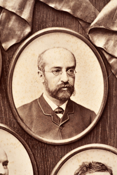 Pośrodku prostokątnego pola o zaokrąglonych narożnikach większa fotografia przyozdobiona wstęgą i gałązkami wawrzynu. Dookoła szesnaście symetrycznie rozmieszczonych mniejszych fotografii. W prawym górnym rogu dr Wacław Lasocki, naczelny lekarz kolei nadwiślańskiej w latach 1875 - 78. W polu ramy w górnej części medalion z płaskorzeźbionym popiersiem z profilu. Przedstawia Leopolda Kronenberga - ojca. Po bokach dwia półleżące aniołki trzymają wieniec laurowy. Po lewej stronie ramy postać robotnika Poleszuka, po prawej pracownika warsztatów kolejowych. W dolnej części front lokomotywy i złoty żeton uprawniający do bezpłatnej jazdy pociągiem I klasy. Fotografia naklejona na tekturę, w dolnej części u dołu sucha pieczęć zakładu fotograficznego.