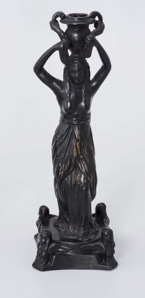 Świecznik figuralny z kariatydą; cokół czworoboczny górą przechodzący w koło, w narożach figurki sfinksów, na nim stojąca postać kobiety z wazonem na głowie, który podtrzymuje uniesionymi rękami. Kobieta do pasa naga, ubrana jest w fałdzistą szatę przwiązaną u góry, na głowie chusta opadajaca na ramiona. Całość malowana lakierem na czarno.
