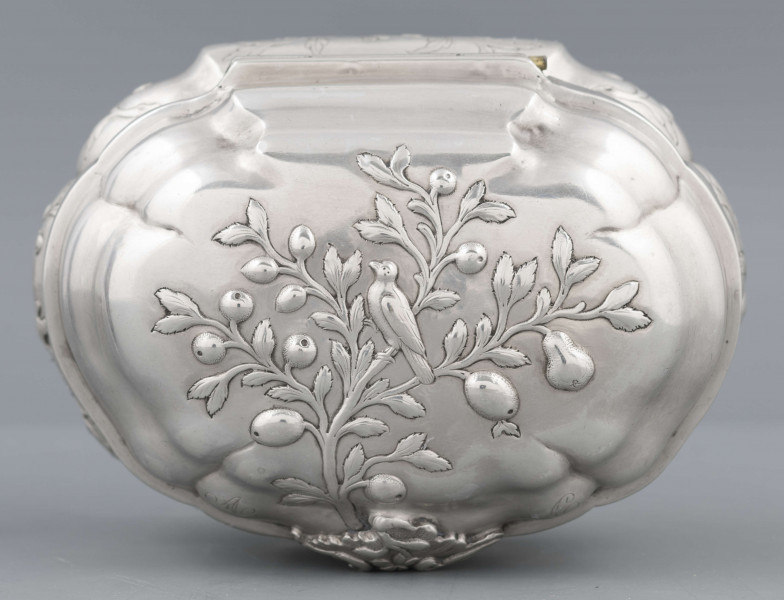 S/Mt/641/ML - Cukiernica owolidalna z zamykanym wiekiem, profilowana, na niskim cokole o falistym wykroju brzegu. Zdobiona repusowaną dekoracją stosującą w różnych wariantach motyw gałązki z kwiatami i owocami, na której siedzi ptak. W przedniej ściance otwór na kluczyk, powyżej w wieczku nałożony ornament maskujący zaczep zamka (brak), po lego bokach grawerowane inicjały 