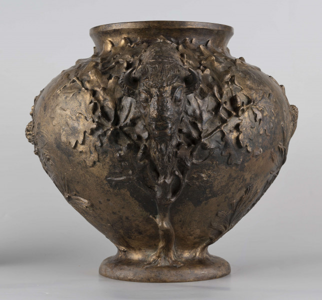 S/Mt/427/ML - Waza w kształcie spłaszczonej kuli, z niską, kolistą stopą i krótką szyją o lekko wywiniętym brzegu. Powierzchnia zdobiona reliefem: po bokach dwa uchwyty w formie głów żubrów, wokół nich gałązki dębu, pośrodku korpusu gałązki sosny z szyszkami wyrastające ze stopy. U góry na brzuścu rytowany napis majuskułą 